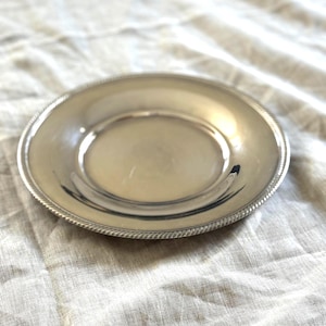 May include: A small, round, silver-colored plate with a decorative rope-like border. The plate has a slightly concave center and reflects the light. The plate is likely made of metal and is suitable for serving small portions.