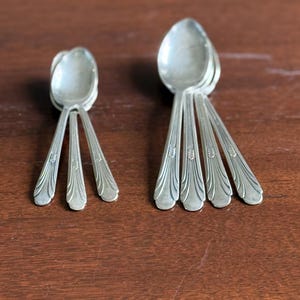 May include: A collection of vintage silver spoons arranged in two groups. The spoons have a classic design with detailed handles and rounded bowls. The spoons are displayed on a dark wooden surface, creating a contrast with the silver.