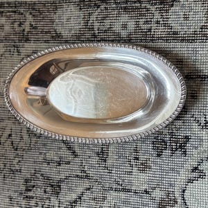 May include: Oval silver-plated serving tray with a rope-like border. The tray has a polished, reflective surface and a shallow, elongated center. The tray is designed for serving food or displaying items.