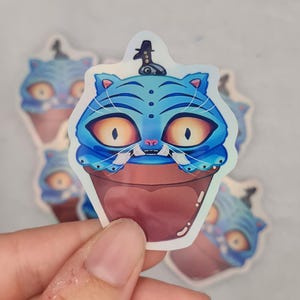 Peut inclure: Autocollant holographique représentant une créature bleue ressemblant à un chat avec de grands yeux jaunes, un petit chapeau et un corps en forme de cupcake. L'autocollant a une base marron et un dessus bleu, avec un motif à rayures blanches et noires. Plusieurs autocollants sont visibles.
