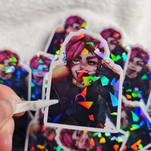 May include: A holographic sticker featuring a cartoon character with pink hair, a black shirt, and a mischievous grin. The character is partially obscured by holographic glitter.