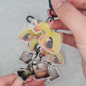 Könnte beinhalten: Eine Sammlung klarer Acryl-Schlüsselanhänger mit Cartoon-Charakteren. Die Schlüsselanhänger haben einen schwarzen Verschluss und einen schwarzen Ring. Die Charaktere tragen gelbe und schwarze Outfits. Die Schlüsselanhänger werden von einer Hand gehalten.