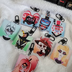 Może przedstawiać: Osiem kolorowych breloczków z postaciami z kreskówek z czarnymi metalowymi klamrami. Postacie pochodzą z serialu animowanego Helluva Boss. Postacie obejmują: Loona, Moxxie, Millie, Blitzo, Stolas, Verosika i postać z czerwonymi włosami z czarnym okiem.