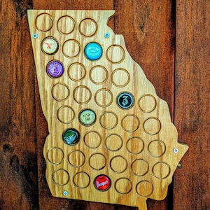 May include: A wooden wall-mounted bottle cap holder shaped like the state of Georgia. The holder has 48 slots for bottle caps and features a few bottle caps already in place.