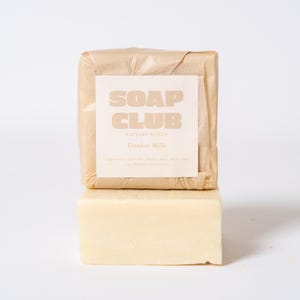 May include: Two rectangular bars of soap stacked vertically. The top bar is wrapped in light brown paper with a white label featuring the text SOAP CLUB and Donkey Milk. The bottom bar is a solid, pale cream-colored soap with a smooth, matte finish.