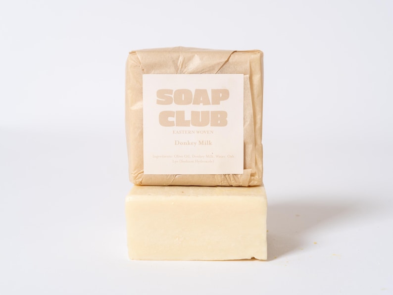 May include: Two rectangular bars of soap stacked vertically. The top bar is wrapped in light brown paper with a white label featuring the text SOAP CLUB and Donkey Milk. The bottom bar is a solid, pale cream-colored soap with a smooth, matte finish.
