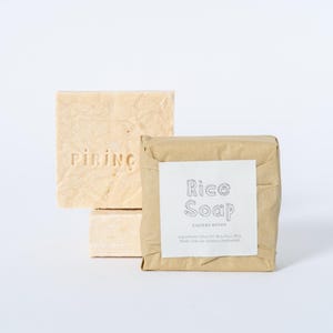 May include: Two rectangular bars of light beige soap. One bar is unwrapped and features the embossed text PIRING. The second bar is wrapped in brown paper with a white label that reads Rice Soap and includes a list of ingredients in small text.