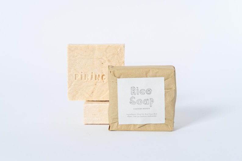 May include: Two rectangular bars of light beige soap. One bar is unwrapped and features the embossed text PIRING. The second bar is wrapped in brown paper with a white label that reads Rice Soap and includes a list of ingredients in small text.