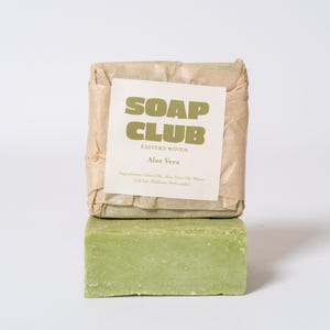 May include: A rectangular bar of light green soap stacked beneath another bar wrapped in tan paper. The paper label features the text SOAP CLUB, EASTERN WOVEN, Aloe Vera, and a list of ingredients including Olive Oil, Aloe Vera Oil, Water, and Lye.