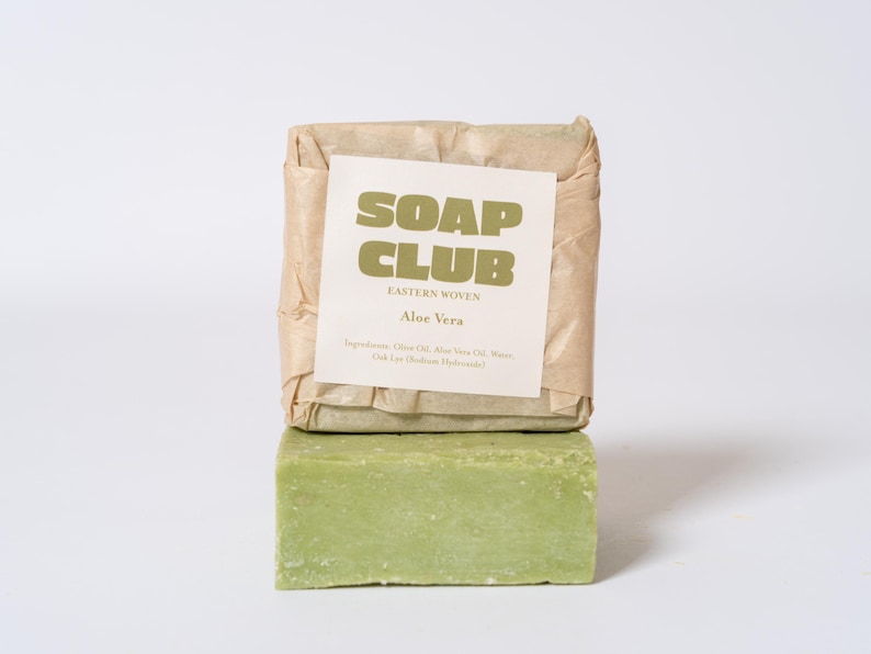 May include: A rectangular bar of light green soap stacked beneath another bar wrapped in tan paper. The paper label features the text SOAP CLUB, EASTERN WOVEN, Aloe Vera, and a list of ingredients including Olive Oil, Aloe Vera Oil, Water, and Lye.