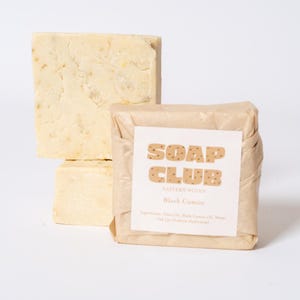 May include: Two rectangular bars of off-white soap, one unwrapped and one wrapped in tan paper. The wrapped bar features a white label with the text SOAP CLUB, EASTERN WOVEN, and Black Cumin, along with a list of ingredients printed in black.