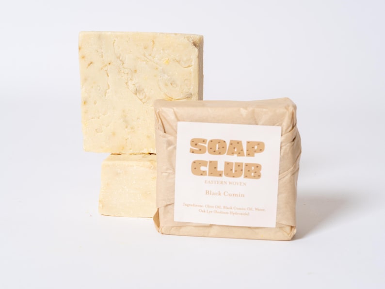 May include: Two rectangular bars of off-white soap, one unwrapped and one wrapped in tan paper. The wrapped bar features a white label with the text SOAP CLUB, EASTERN WOVEN, and Black Cumin, along with a list of ingredients printed in black.