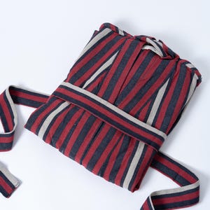 May include: Folded striped robe in red, navy, and beige. The robe features vertical stripes and a matching belt. The fabric appears to be soft and comfortable. The robe is folded neatly, ready for use.