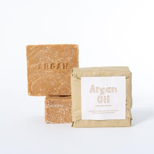 May include: Two square bars of light brown argan oil soap with the word ARGAN embossed on the front. One bar is wrapped in tan paper with a white label featuring the text Argan Oil and a list of ingredients printed in black ink.
