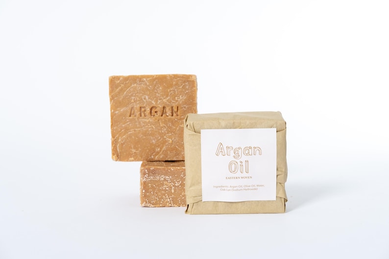 May include: Two square bars of light brown argan oil soap with the word ARGAN embossed on the front. One bar is wrapped in tan paper with a white label featuring the text Argan Oil and a list of ingredients printed in black ink.