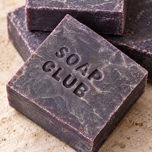 May include: A stack of square, dark purple bars of soap with a textured, marbled surface. The top bar features the embossed text SOAP CLUB in a clean, sans-serif font. Each bar has a solid, dense appearance with subtle variations in color tone.