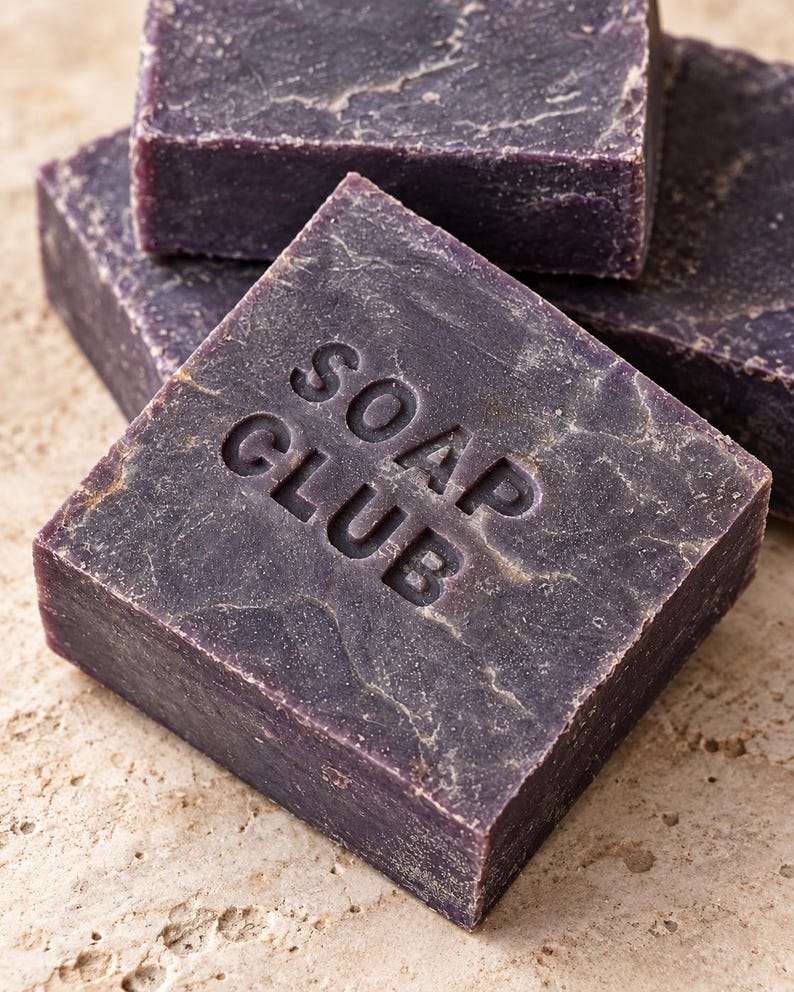 May include: A stack of square, dark purple bars of soap with a textured, marbled surface. The top bar features the embossed text SOAP CLUB in a clean, sans-serif font. Each bar has a solid, dense appearance with subtle variations in color tone.
