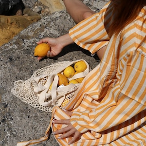 May include: A person wearing a yellow and white striped shirt sits on a rock with a mesh bag filled with lemons.