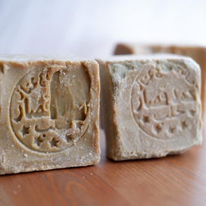 May include: Close-up of three rectangular, light brown soaps with embossed Arabic script and star designs. The soaps are on a wooden surface, with the front two in sharp focus and the third slightly blurred in the background.