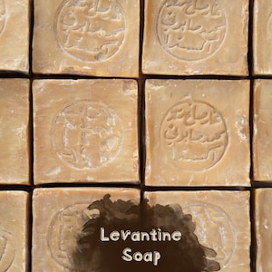 May include: A grid of twelve square, light brown bars of soap arranged in four rows and three columns. Each bar features a circular stamp with Arabic text embossed in the center. The words Levantine Soap are printed in white over a dark brown blotch.