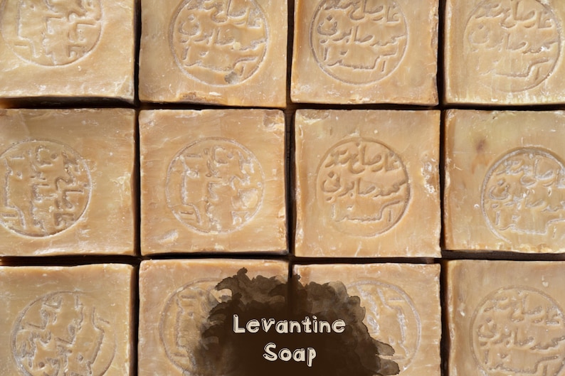 May include: A grid of twelve square, light brown bars of soap arranged in four rows and three columns. Each bar features a circular stamp with Arabic text embossed in the center. The words Levantine Soap are printed in white over a dark brown blotch.