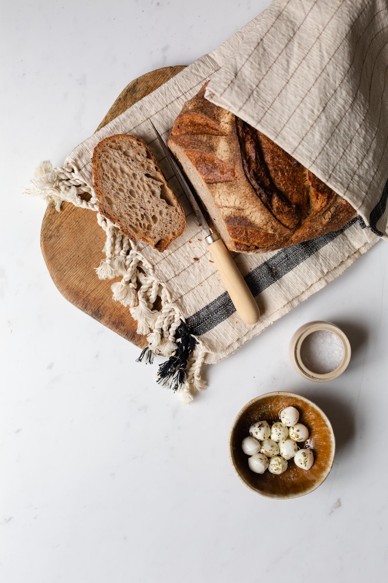 Peut inclure: Une miche de pain au levain sur une planche &agrave; d&eacute;couper en bois avec un couteau et un bol de boulettes de mozzarella. Le pain est partiellement tranch&eacute; et le fromage est saupoudr&eacute; d'herbes.