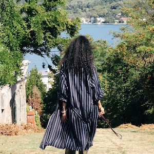 May include: A person wearing a long, striped, navy blue and white shirt dress with a long, dark brown leather strap hanging from their wrist. They are walking on a grassy path with a body of water in the background.