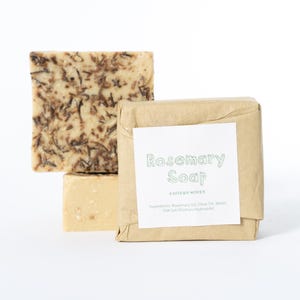 May include: Three square bars of soap. One bar is unwrapped, showing a beige color with dark brown herb flecks. Another bar is wrapped in brown paper with a white label reading Rosemary Soap, Eastern Woven, and a list of ingredients.