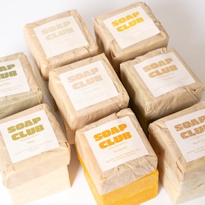 May include: Eight square bars of soap wrapped in brown paper, each featuring a white label with the text SOAP CLUB in bold yellow or green letters. The bars are arranged in a grid, showcasing various colors including pale yellow, orange, and green.