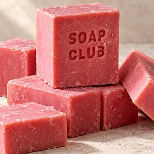 May include: A collection of solid, square-shaped pink soap bars with a textured, matte surface. The central, upright bar features the embossed text "SOAP CLUB" on its front face. The bars are arranged in a stacked formation with varying angles.