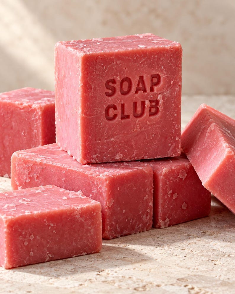 May include: A collection of solid, square-shaped pink soap bars with a textured, matte surface. The central, upright bar features the embossed text "SOAP CLUB" on its front face. The bars are arranged in a stacked formation with varying angles.