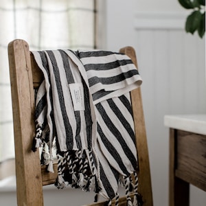 Peut inclure: Une serviette ray&eacute;e noire et blanche avec des franges est accroch&eacute;e &agrave; une chaise en bois. La serviette porte une petite &eacute;tiquette indiquant "Eastern Weavers" et "Made in Turkey".