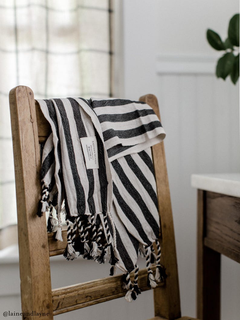 Peut inclure: Une serviette ray&eacute;e noire et blanche avec des franges est accroch&eacute;e &agrave; une chaise en bois. La serviette porte une petite &eacute;tiquette indiquant "Eastern Weavers" et "Made in Turkey".