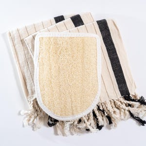 Op de afbeelding: Een wit-zwarte gestreepte handdoek met een natuurlijke loofah spons bevestigd. De handdoek heeft franjes.