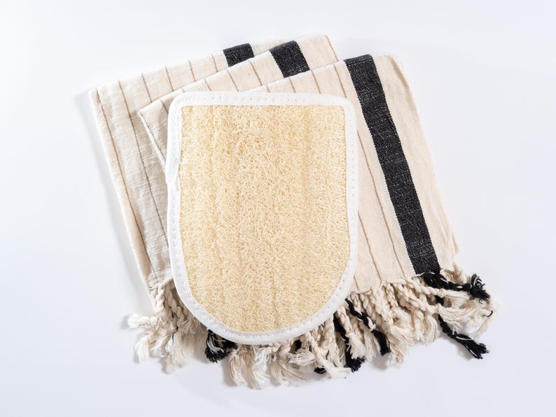 Op de afbeelding: Een wit-zwarte gestreepte handdoek met een natuurlijke loofah spons bevestigd. De handdoek heeft franjes.