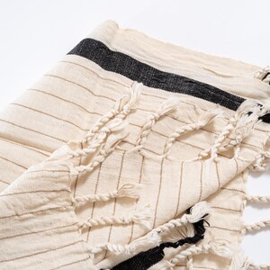May include: A white cotton towel with black stripes and fringe. The towel is folded in half and the fringe is visible on both sides.