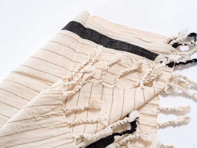 May include: A white cotton towel with black stripes and fringe. The towel is folded in half and the fringe is visible on both sides.