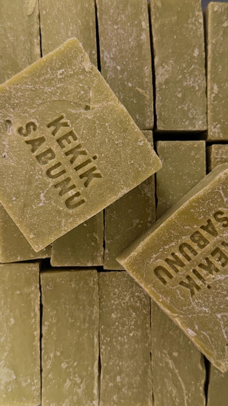 May include: A stack of rectangular olive green bars of soap. Each bar has a textured, matte surface and is stamped with the text KEKIK SABUNU in a clear, sans-serif font. The bars are arranged in a grid-like pattern, showing their edges and faces.
