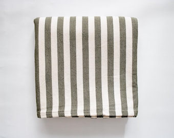 Op de afbeelding: Een rechthoekige textiele plaid met een herhalend verticaal streeppatroon in afwisselende kleuren olijfgroen en gebroken wit. De stof heeft een zichtbare geweven textuur en is netjes opgevouwen in een compacte, vierkante vorm.