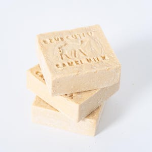 Puede incluir: Una pila de tres barras de jabón cuadradas de color beige claro. La barra superior tiene las palabras "EVE CUTLO" y "CAMEL MILK" grabadas, junto con la silueta de un camello. Fondo blanco.
