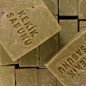 May include: A stack of rectangular olive green bars of soap. Each bar has a textured, matte surface and is stamped with the text KEKIK SABUNU in a clear, sans-serif font. The bars are arranged in a grid-like pattern, showing their edges and faces.