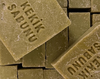 Può includere: Una pila di saponette rettangolari color verde oliva con una superficie ruvida e opaca. Due delle saponette sono marchiate con il testo KEKIK SABUNU in un carattere sans-serif chiaro, mentre le altre rimangono semplici e prive di decorazioni.