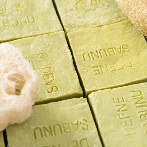 May include: Close-up of multiple green bars of soap with the text "DEFNE SABUNU" embossed on each bar. A natural loofah sponge is in the foreground.