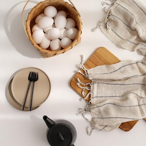 Peut inclure: Un panier en osier blanc rempli d'&oelig;ufs blancs, une th&eacute;i&egrave;re noire, deux assiettes beiges avec une fourchette noire et deux serviettes ray&eacute;es sur une surface blanche.