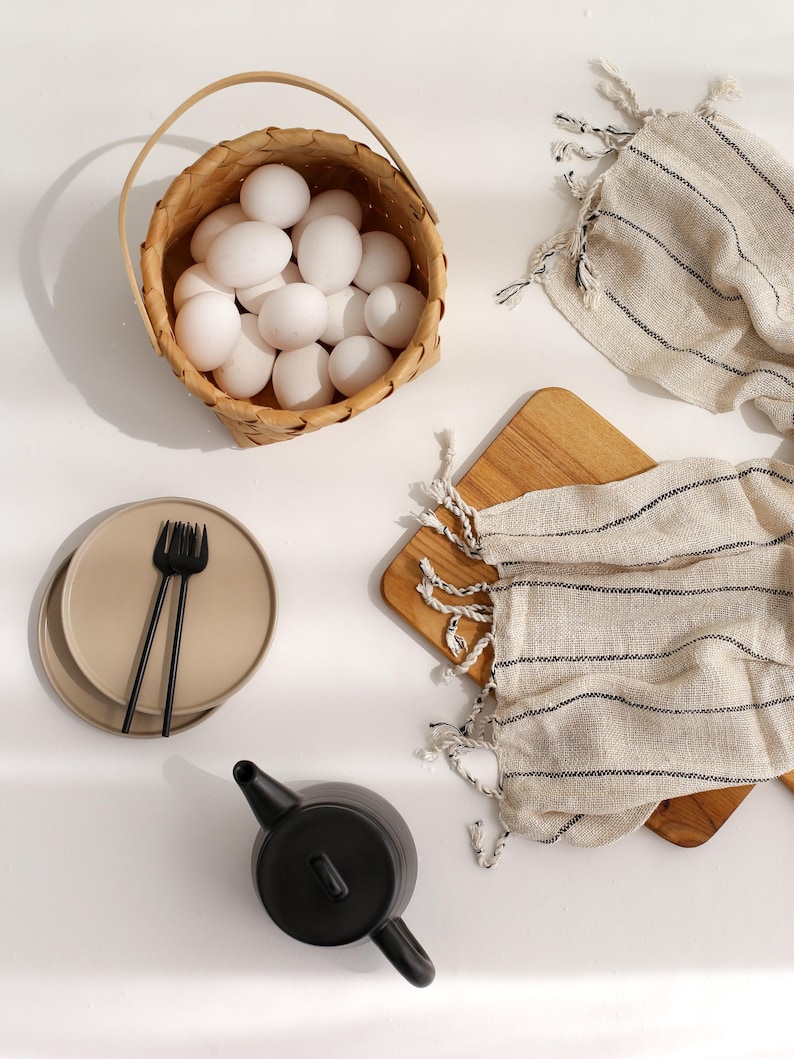 Peut inclure: Un panier en osier blanc rempli d'&oelig;ufs blancs, une th&eacute;i&egrave;re noire, deux assiettes beiges avec une fourchette noire et deux serviettes ray&eacute;es sur une surface blanche.