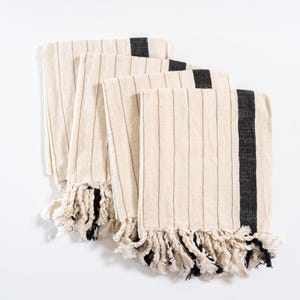 May include: Four cream-colored kitchen towels with black stripes and fringe. The towels are folded and stacked on a white surface.