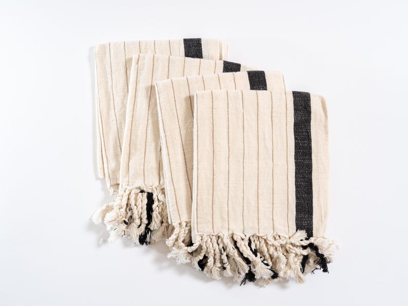 May include: Four cream-colored kitchen towels with black stripes and fringe. The towels are folded and stacked on a white surface.