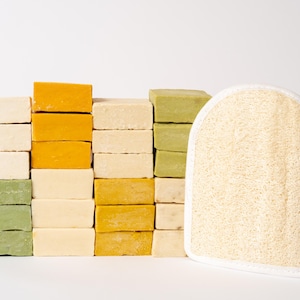 May include: A collection of rectangular soap bars in shades of cream, yellow, and sage green stacked in columns, accompanied by a single beige loofah exfoliating mitt with a white fabric border and a rounded top edge.