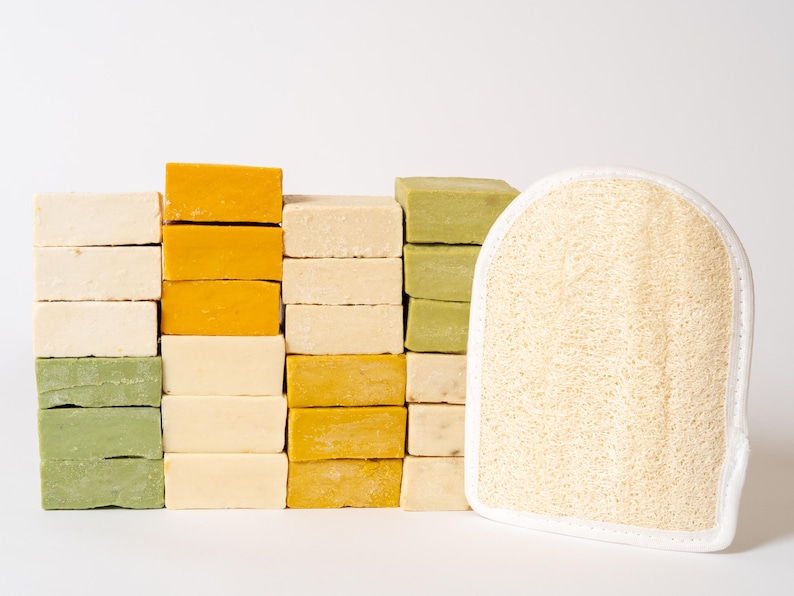 May include: A collection of rectangular soap bars in shades of cream, yellow, and sage green stacked in columns, accompanied by a single beige loofah exfoliating mitt with a white fabric border and a rounded top edge.