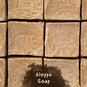 May include: A grid of square, light brown Aleppo soap bars, each featuring a circular stamped emblem with Arabic calligraphy and small stars. A dark brown, irregular watercolor-style patch is overlaid at the bottom, displaying the text Aleppo Soap.